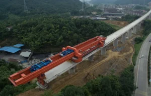 Máquina lanzadora de vigas sobre viaducto ferroviario elevado en construcción, rodeado de colinas verdes y zona urbana