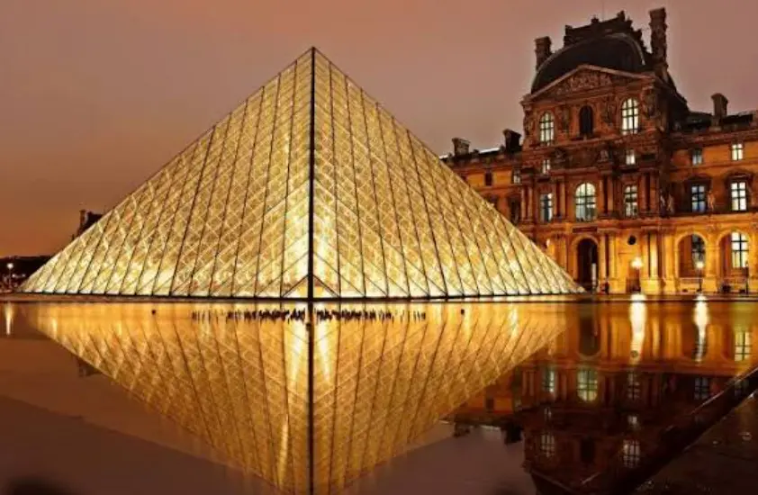 Pirámide de cristal del Museo del Louvre iluminada al atardecer frente al palacio histórico de París.
